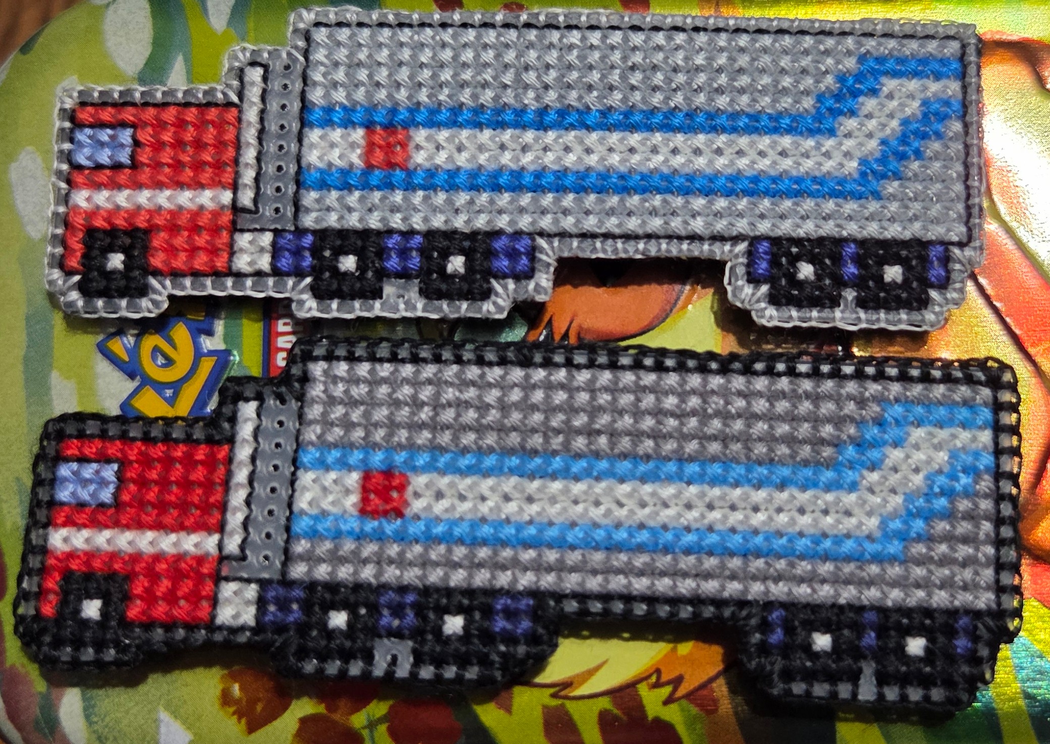 Two cross stitch pieces of a semi truck with trailer, based on the character Optimus Prime's design. Truck is red, while the trailer is grey, blue, and white. The colors vary slightly between the two, but are otherwise the same.