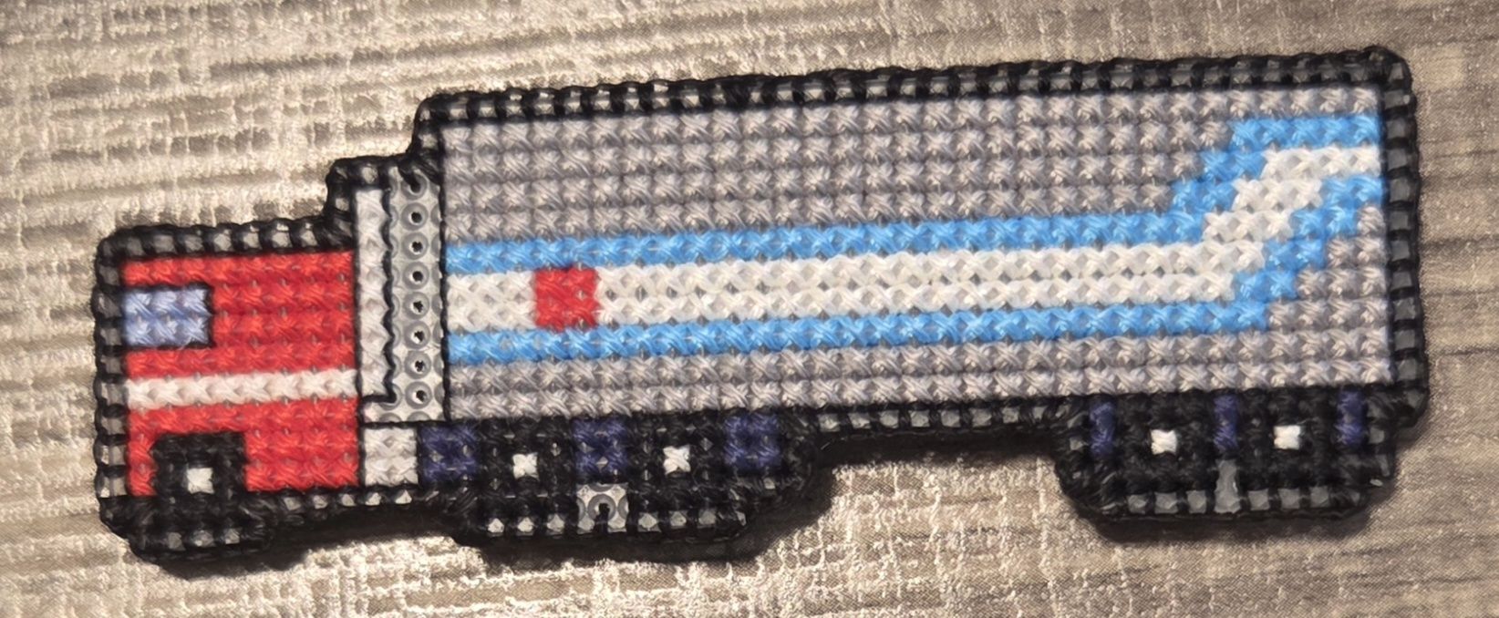 Cross stitch piece of a semi truck with trailer, based on the character Optimus Prime's design. Truck is red, while the trailer is grey, blue, and white.