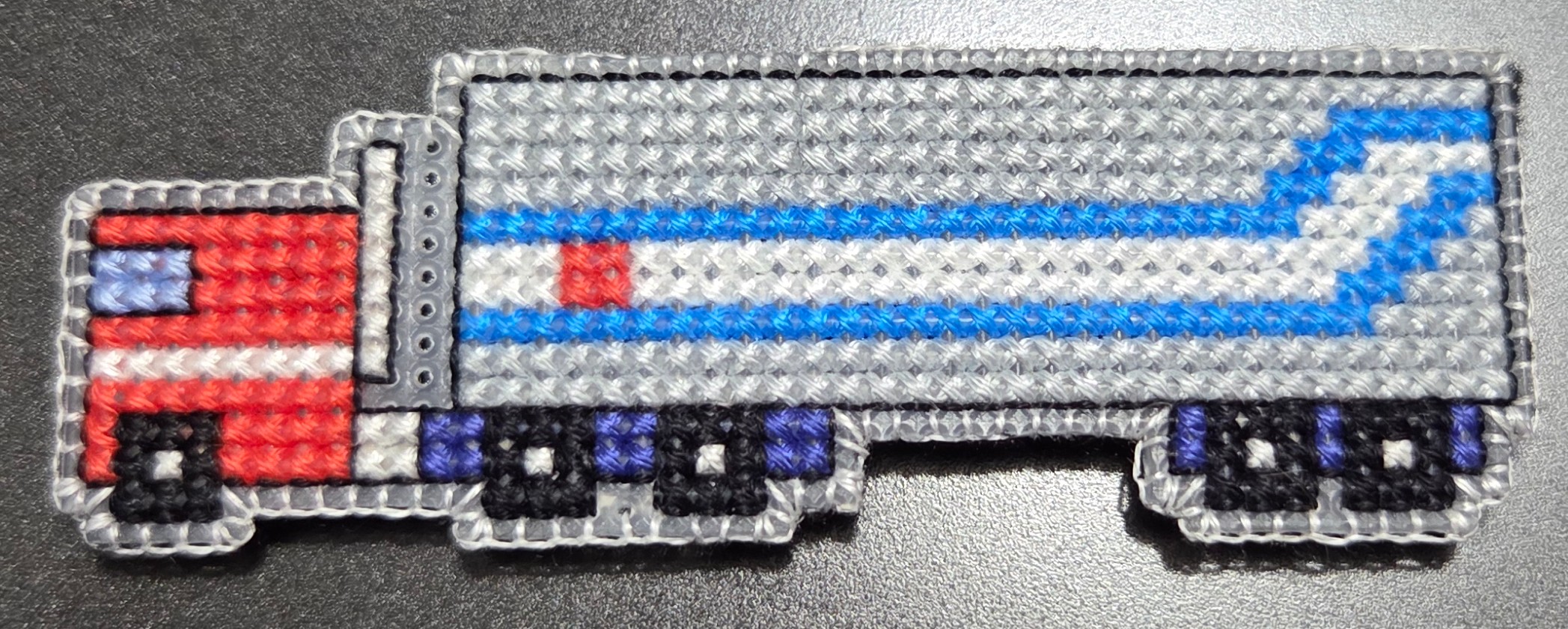 A different cross stitch piece of a semi truck with trailer, based on the character Optimus Prime's design. Truck is red, while the trailer is grey, blue, and white.