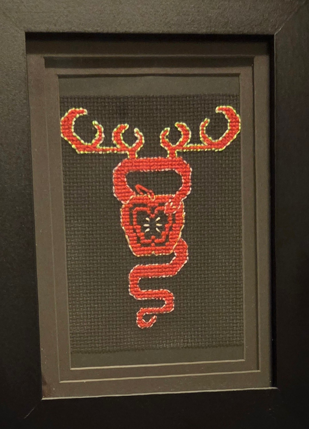 Framed cross stitch. The image is in red. It depicts an apple, a snake curling around the apple, and antlers coming off the snake towards the top.
